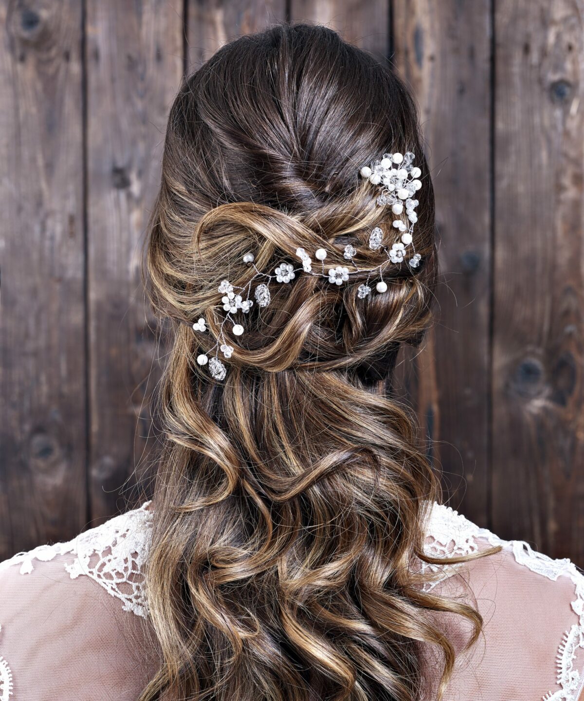 Dunkelhaarige Braut mit festlicher Frisur trägt einen Haardraht mit Blumen und Perlen von CELEBRIDE Trisa Accessoires, vor dunkler Holzwand.